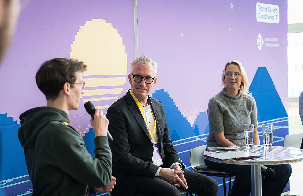 Foto einer Paneldiskussion: Eine junge Person hält das Mikro in der Hand, die anderen hören ihm zu.