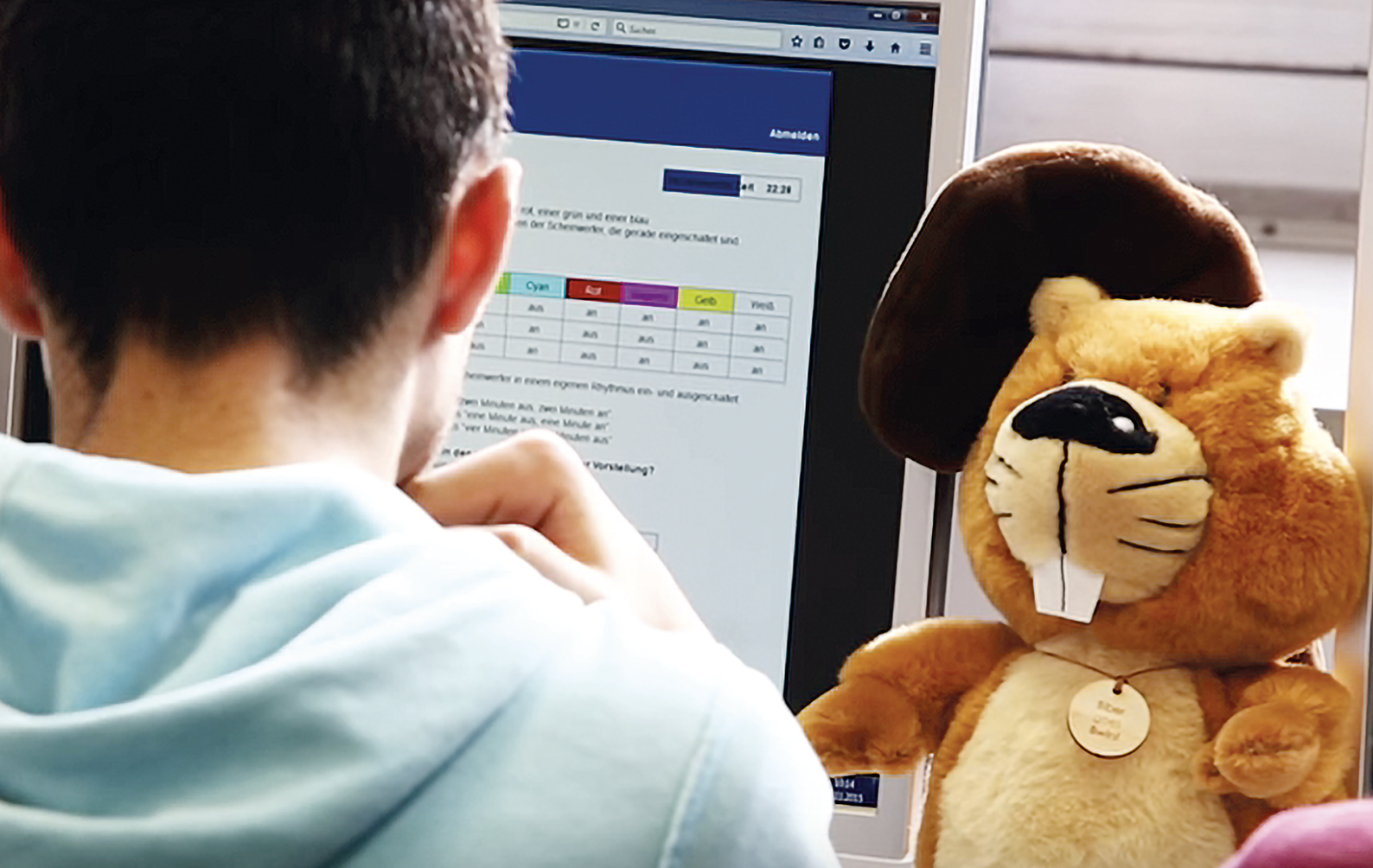 Ein Kind sitzt am Computer und löst eine der Wettbewerbsaufgaben, daneben sitzt ein Plüsch-Biber.