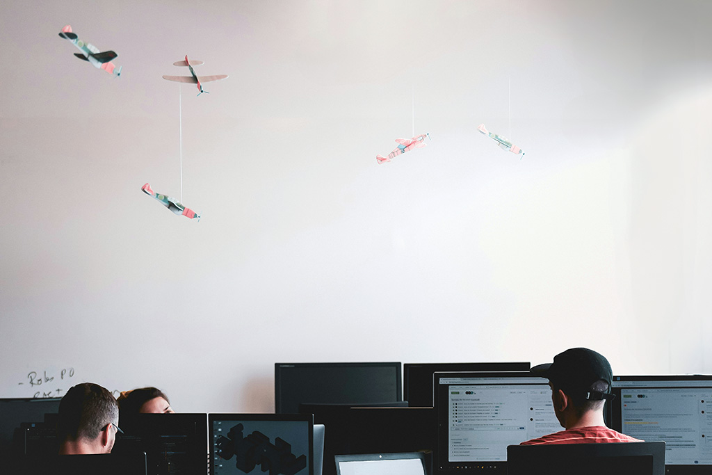 Blick in ein Entwicklerbüro: Mehrere Menschen sitzen an Rechnern, über ihnen hängen bunte Flugzeuge aus Papier