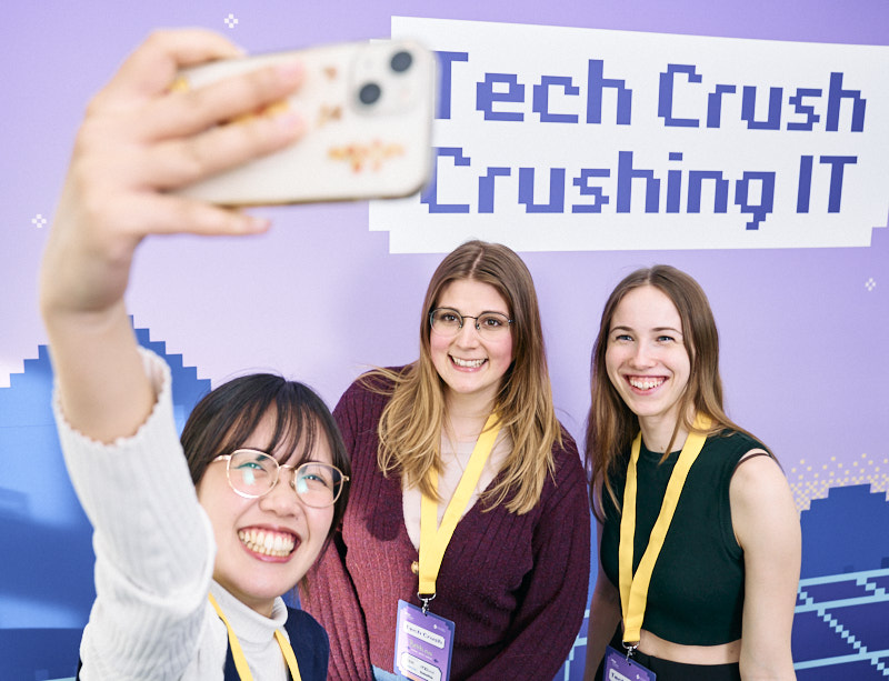 drei junge und fröhliche Frauen, welche vor einem Tech Crush Aufsteller ein Selfie machen