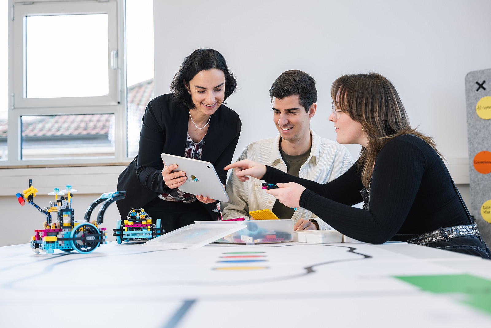 Drei Menschen im Gespräch, eine von ihnen hält ein iPad, auf das eine andere Person zeigt., auf dem Schreibtisch neben ihnen stehen Roboter aus Plastikbauklötzen.
