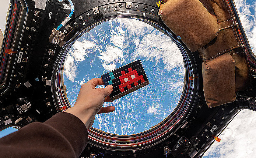 Aufnahme aus einem echten Raumschiff: Eine Hand hält ein Mosaik des im Text erwähnten Künstlers mit einem Space Invader vor ein rundes Fenster. Dahinter ist die Erde von oben zu sehen. 