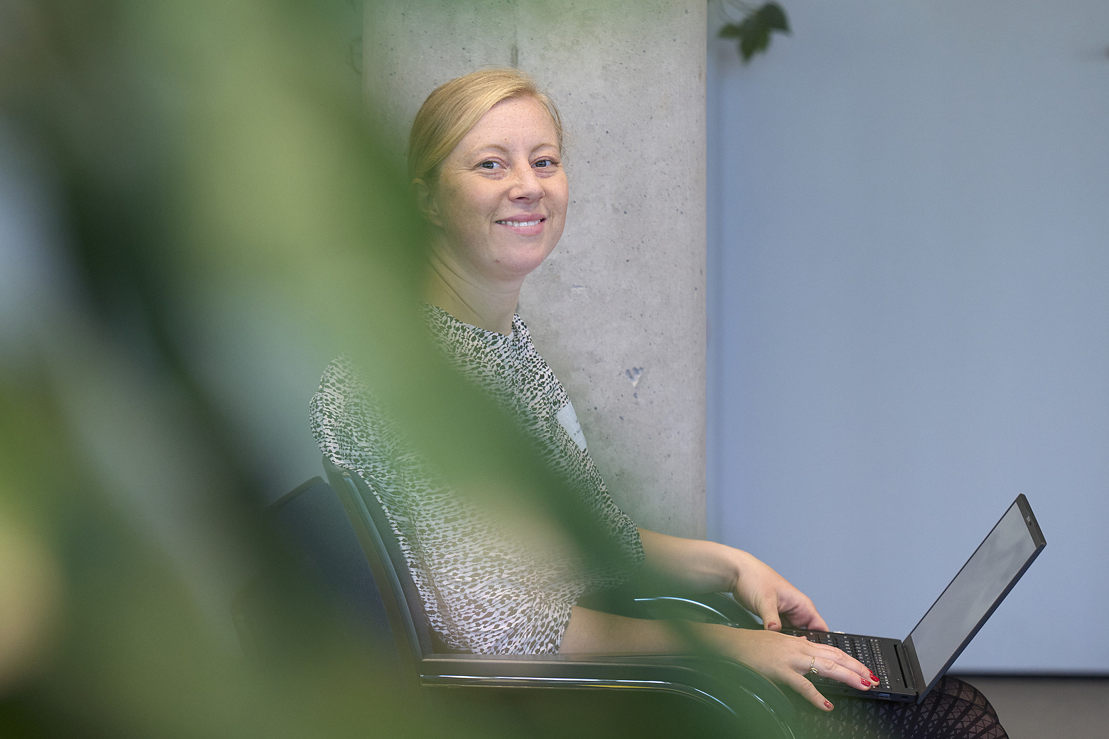 Eine Frau sitzt auf einem Stuhl und hat ihren geöffneten Laptop auf dem Schoß, sie lächelt in die Kamera