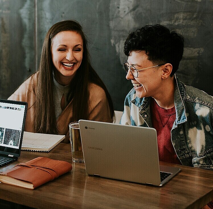 Zwei junge Frauen mit aufgeklappten Laptops in einem Meetingraum, sie lachen.