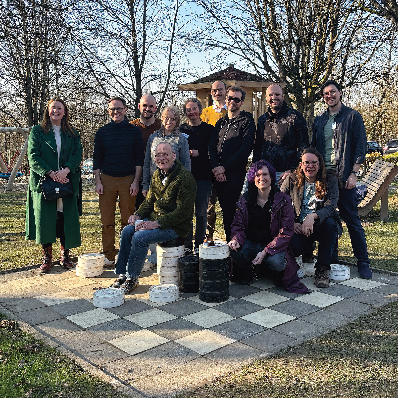 Eine Gruppe lächelnder Menschen in einem Park in der Abendsonne, sie stehen auf einem überdimensionalen Schachbrett und lehnen sich zum Teil an die Spielfiguren.