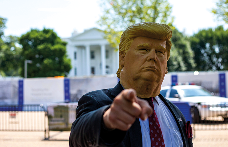 Eine Person, die eine Gummimaske mit dem Gesicht von Donald Trump trägt, steht vor dem Weißen Haus und zeigt mit dem Zeigefinger in die Kamera