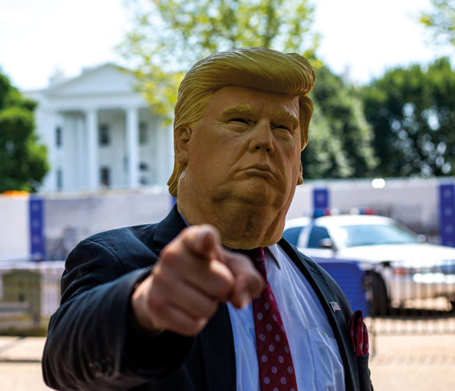 Eine Person, die eine Gummimaske mit dem Gesicht von Donald Trump trägt, steht vor dem Weißen Haus und zeigt mit dem Zeigefinger in die Kamera