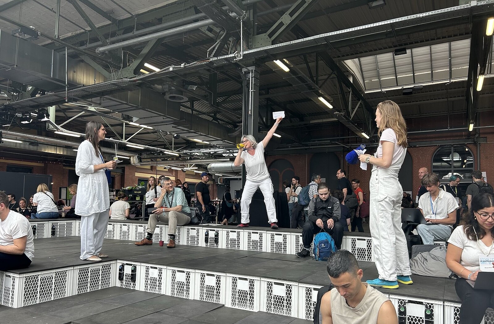 Drei Frauen in Weiß gekleidet stehen in der Meetup-Zone der re:publica-Konferenz. Eine von ihnen hat die Hand zum Himmel gestreckt und ruft etwas.