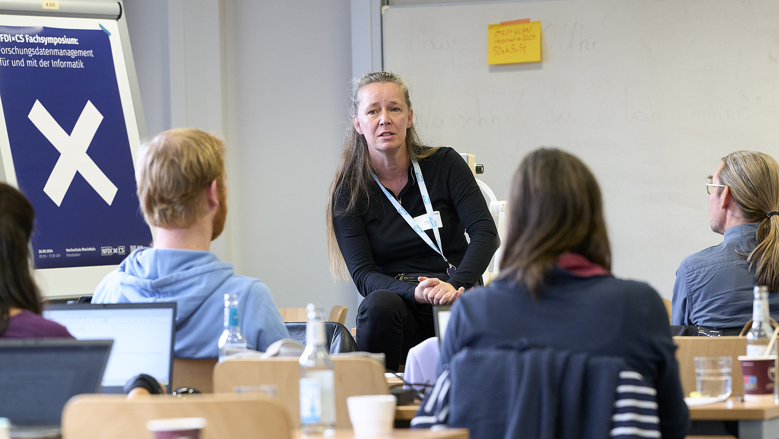 Szene in einem Workshopraum: Ulrike Lucke lehnt auf einem Schreibtisch und ist im Gespräch mit Teilnehmenden