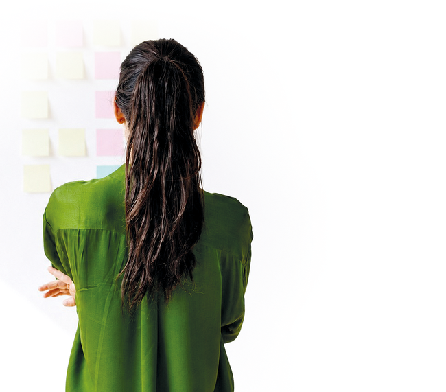 Foto einer jungen Frau von hinten, sie blickt auf eine Wand mit Post-its und hat die Arme verschränkt, als würde sie nachdenken