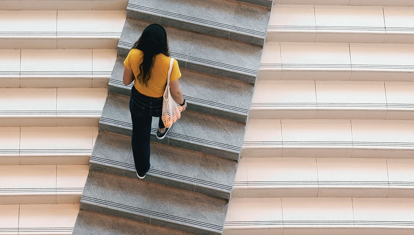 Foto einer Person, die eine Treppe emporsteigt. Sie trägt einen Beutel mit Orangen und ein hellgelbes T-Shirt.