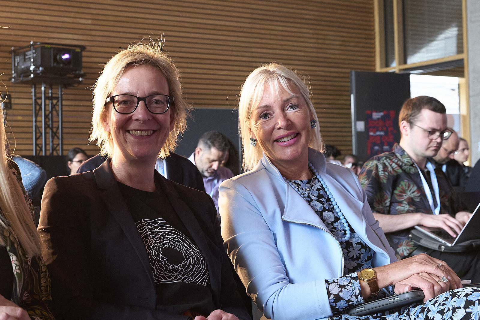 Christine Regitz und Kristina Sinemus sitzen im Publikum und lächeln in die Kamera, Christine Regitz trägt ein T-Shirt aus der GI-Merch-Kollektion