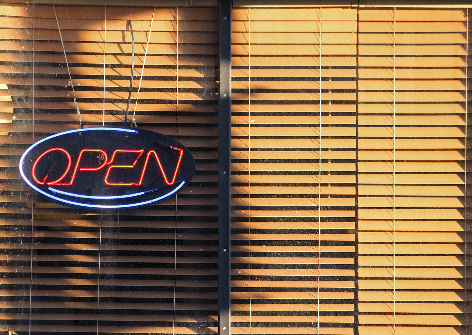 Ein Blick auf ein Fenster eines Restaurants, im Fenster hängt ein Neonschild, auf dem „OPEN“ steht, die Jalousien verdecken aber den Blick hinein