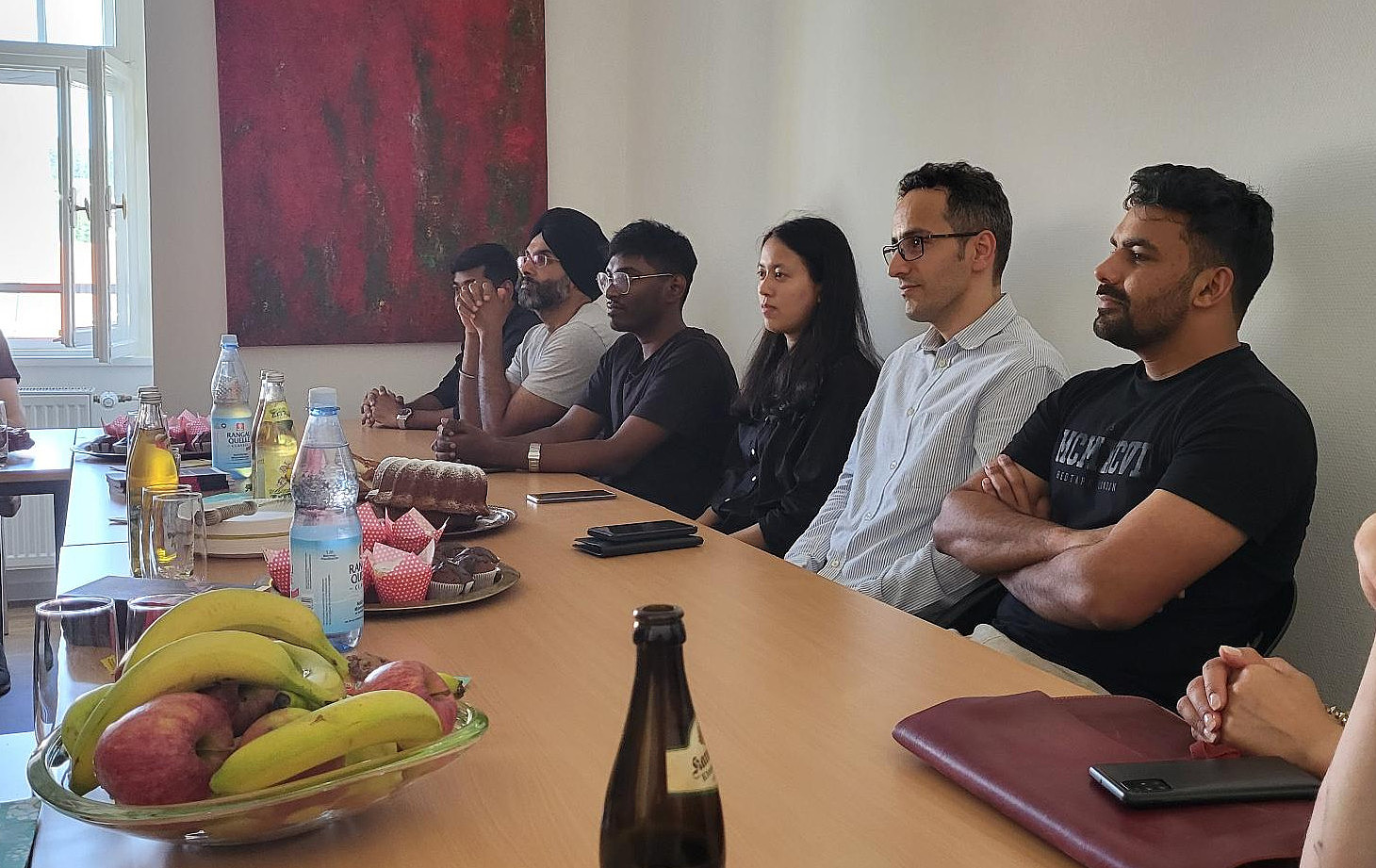 Eine Gruppe Studierender sitzt in einem anderen Seminarraum, vor ihnen Kuchen und Getränke. Sie scheinen jemandem aufmerksam zuzuhören.