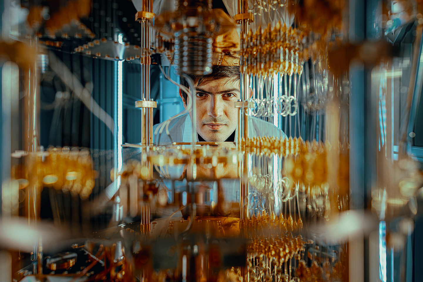 Ein Forscher, der durch Bauteile eines Quantencomputers hindurch in die Kamera blickt 