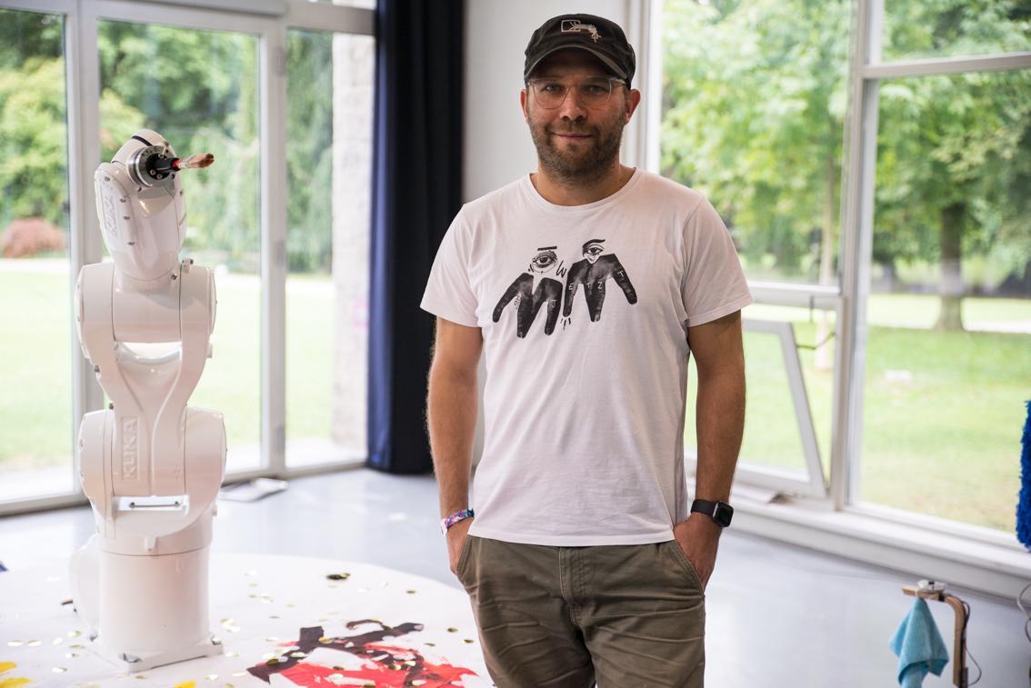 Porträt von Markus Schubert, er trägt eine Baseballcap und steht nebem einem Industrieroboter mit Greifarm