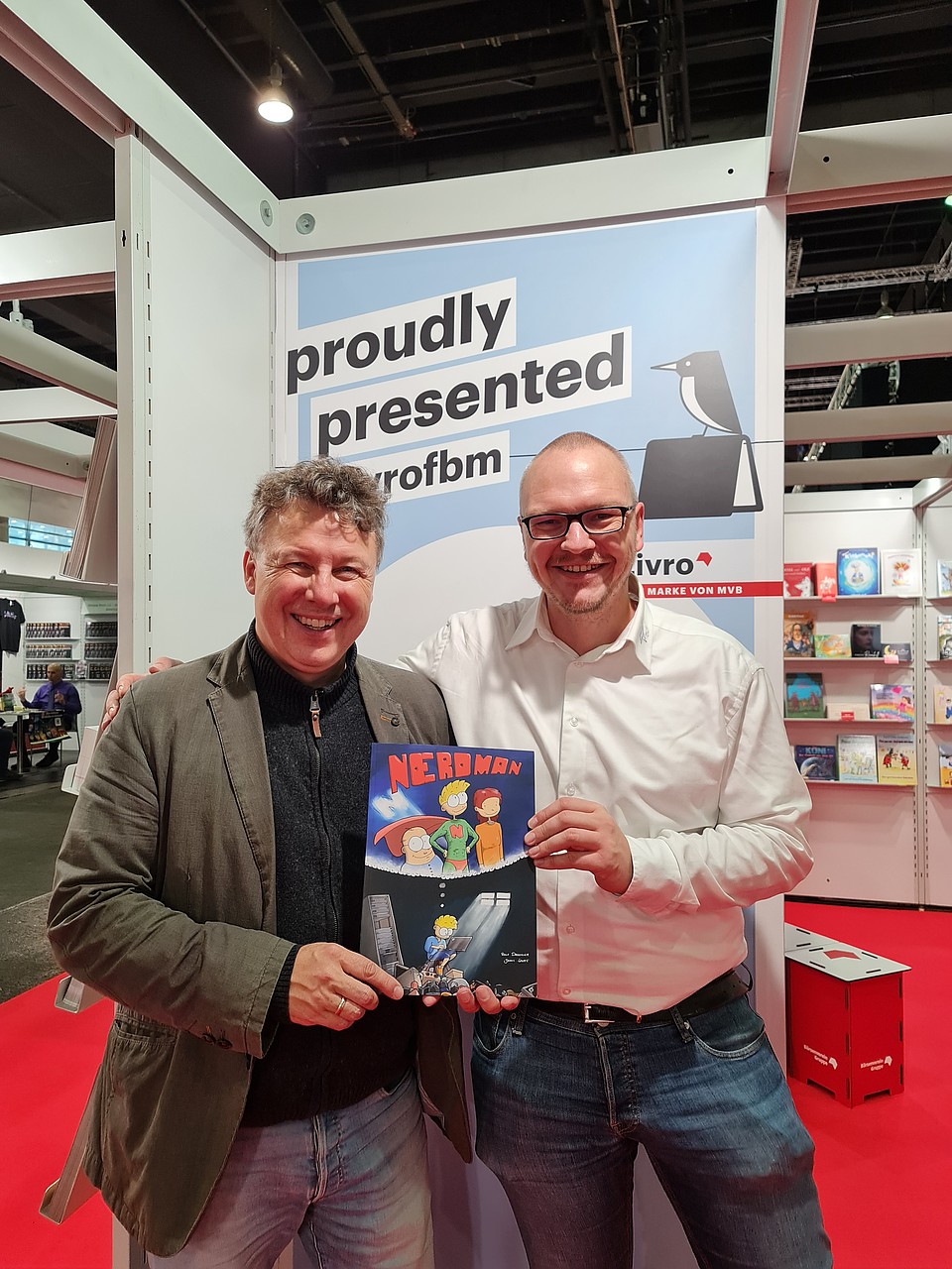 Rolf Drechsler und Jannis Stoppe auf der Frankfurter Buchmesse: Sie halten stolz ihr neu veröffentlichtes Buch in der Hand, auf dessen Cover eine nerdige Superheldenfigur abgebildet ist.